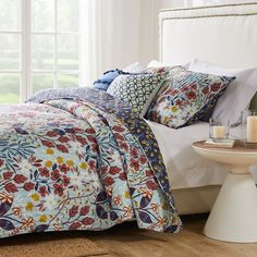 a bed with white sheets and colorful flowers on it, next to a night stand