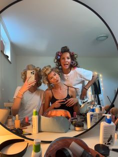 three women sitting in front of a mirror with hair dryers and makeup products on the counter