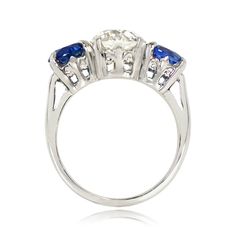 This antique three-stone engagement ring centers a 2.04 carat old European cut diamond, J color and VS1 clarity, set in prongs. The diamond is accented by two round cut sapphires with a combined weight of 2.56 carats, also set in prongs. The handcrafted platinum mounting features a fleur-de-lis motif on the under-gallery. This ring is from the Art Deco era, circa 1930.
The old European diamond measures approximately 7.83mm x 7.78mm x 5.19mm. The sapphires measure approximately 6.31mm x 6.16mm x Art Deco Three Stone Diamond Ring, Art Deco Three Stone Round Ring, Fine Jewelry Three Stone Lab-created Sapphire Ring, Three Stone Lab-created Sapphire Diamond Ring, Sapphire Three Stone Round Cut Diamond Ring, Three Stone Sapphire Round Ring Fine Jewelry, Three Stone Sapphire Diamond Ring With Round Cut, Heirloom Sapphire Three Stone Ring, Vintage Sapphire Three Stone Ring