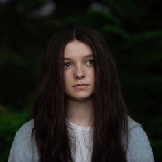 a woman with long brown hair and blue eyes looks into the camera while standing in front of trees