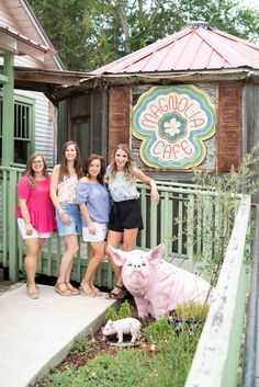 three girls posing in front of a small building with a pig statue on the porch