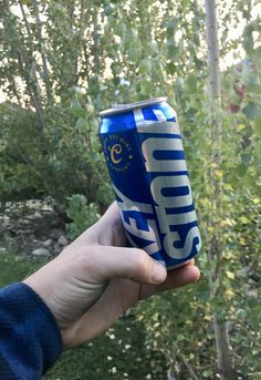 a person holding up a can of soda in front of some trees and bushes with green leaves