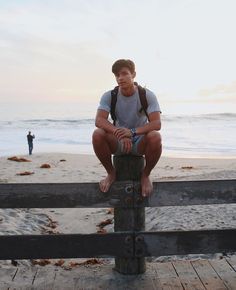 a man sitting on top of a wooden fence next to the ocean and another person standing in the background