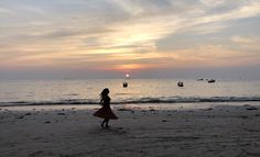 a woman walking on the beach at sunset with boats in the water and clouds overhead