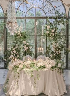 a table with flowers and greenery on it in front of a large glass window