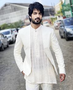 a man with black hair and beard standing in the middle of a street wearing a white suit