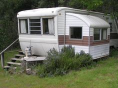 an old camper sits in the grass next to some bushes and trees, with stairs leading up to it