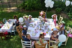 a group of children sitting at a table with balloons and plates on it in the grass