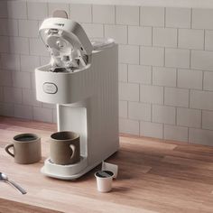 a white coffee maker sitting on top of a wooden counter next to two mugs