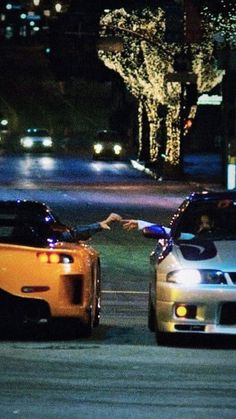 two cars parked next to each other on a street at night with lights in the background