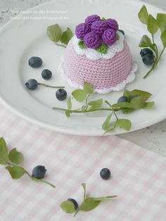 a small crocheted cake on a plate with blueberries and leaves around it