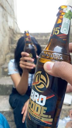 a person holding up a bottle of beer in front of two people sitting on steps