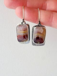 A pair of rounded rectangular Laguna Lace agate pairs are set in fine and sterling silver. Colorful bands of deep red, periwinkle and rose move in wavy stripes through the stones: a beautiful desert sunset palette. A free-moving back layer of sterling silver mirrors the shape of the stone and extends down below it to show the unique texture of silver dust fused to silver. A window cut behind the stones allows you to view the backside of these unusual natural agates. The silver is oxidized to give a beautiful contrast between the stones and the textured metal. These one of a kind earrings measure 1 5/8" in length from the top of the ear wire, and 1/2" wide. Sunset Palette, Beautiful Desert, Desert Sunset, Silver Mirrors, Lace Agate, Move In, Ear Wire, Stone Earrings, Deep Red