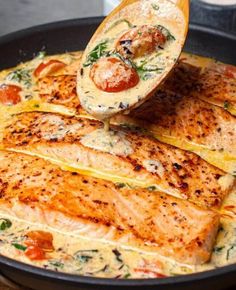 salmon being cooked in a skillet with tomatoes and spinach on the side by a wooden spoon