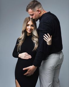 a pregnant woman and man pose for a photo in front of a gray background with their arms around each other