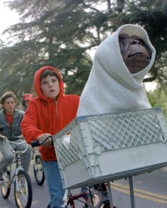 two people riding bikes with a monkey in a hood on the back and another person pushing a bicycle behind them