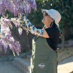 a woman in an apron and hat is picking flowers from a wistery tree