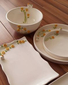 three white dishes with yellow flowers on them sitting on a wooden floor next to a plate and bowl