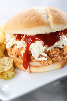 a pulled pork sandwich with cole slaw on a white plate next to potato chips