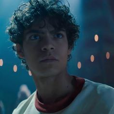 a young man with curly hair stares into the distance while standing in front of lights