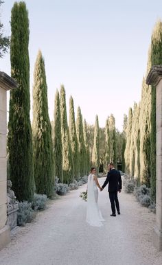 a bride and groom holding hands while walking down a path between two stone pillars with trees in the background