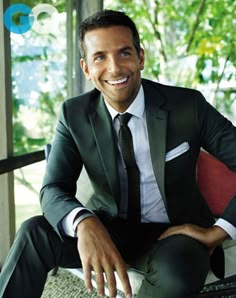 a man in a suit and tie sitting on a chair smiling at the camera with his hands crossed