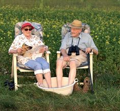 an older couple sitting in chairs on the side of a field with a cat and camera