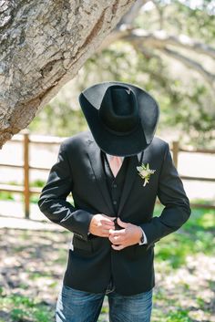 a man wearing a black cowboy hat standing next to a tree in the grass with his hands on his hips