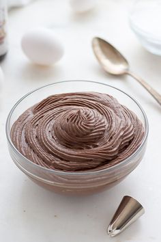 a bowl filled with chocolate frosting next to two spoons and an egg in the background