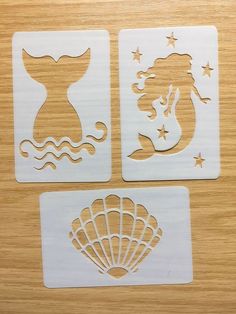 three cutouts of mermaids and seashells on a wooden surface with stars