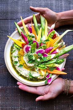 two hands holding a bowl filled with different types of veggies on top of a wooden table