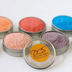 six tins with different colored powdered items in them on a white table top