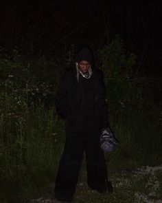 a person standing in the grass at night wearing a black snow suit and holding a handbag