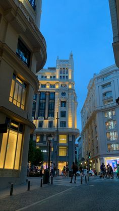 people are walking down the street in front of tall buildings at dusk, with lights on