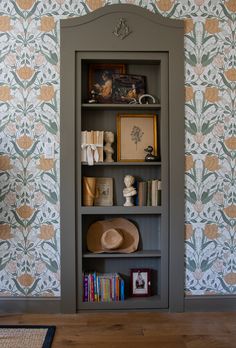 a book shelf with books and hats on it in front of a wallpapered wall