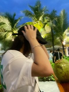 a woman in white shirt and black hat talking on cell phone next to palm trees