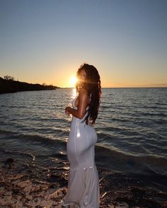 a woman in a white dress standing on the beach at sunset with her back to the camera