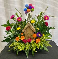 a birdhouse surrounded by colorful flowers and greenery on a black tableclothed surface