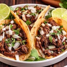 three shredded beef tacos on a plate with limes and cilantro garnishes