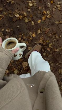 a person holding a cup of coffee in their hand while standing next to leaves on the ground