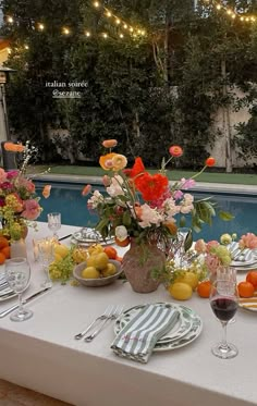 an outdoor table set with flowers, fruit and wine glasses for dinner by the pool