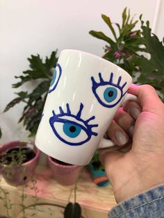 a hand holding a coffee cup with blue eyes on it and plants in the background