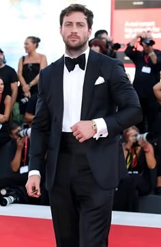 a man in a tuxedo standing on a red carpet with people taking pictures behind him