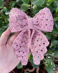 a pink crocheted bow is being held by someone's hand in front of some green leaves