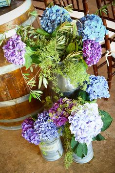 purple and blue flowers sit in buckets next to a wooden barrel on the ground