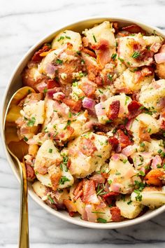 a bowl filled with potatoes and bacon on top of a marble countertop next to a gold spoon