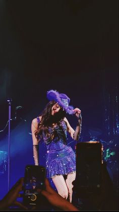 a woman in a purple dress and hat on stage with her hands behind her head