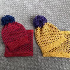 two knitted hats laying next to each other on top of a gray blanket with blue, yellow and red pom - poms