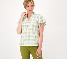 a woman standing in front of a white background wearing green pants and a plaid shirt