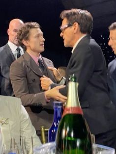 three men in suits are talking to each other at a table with wine bottles and glasses on it